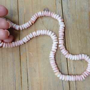 Puede incluir: Un collar de cuentas de ópalo rosa con un cierre de oro. El collar está hecho de pequeñas cuentas redondas que están enhebradas en una cadena fina.