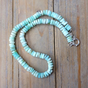 May include: A light blue opal bead necklace with a silver clasp. The necklace is made of round, flat beads that are strung together on a silver chain.