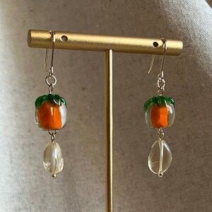 May include: A pair of gold earrings with a clear glass pumpkin charm and a clear quartz stone dangle. The pumpkin charm is orange with green leaves.
