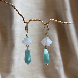 Puede incluir: Un par de pendientes colgantes de oro con un dije en forma de nube blanca y un dije en forma de lágrima verde.