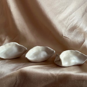 May include: Three white ceramic dumplings, shaped like traditional Chinese dumplings, are arranged in a row on a light brown fabric background.