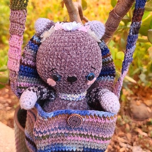 May include: A purple crocheted plush toy with a white bow and sparkly eyes is sitting inside a colorful crocheted bag with a brown button. The bag is hanging from a branch of a green plant.