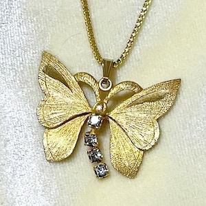 May include: A gold-colored butterfly pendant necklace. The butterfly has textured wings and is adorned with three small, clear gemstones. The necklace chain is also gold-colored and has a delicate, box-style link.