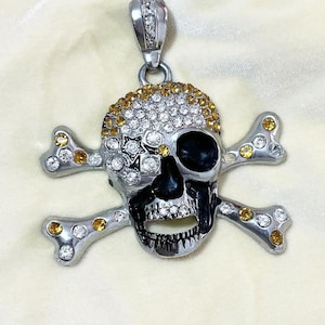 May include: A silver skull and crossbones pendant with clear and yellow rhinestones. The skull has a black eye patch.