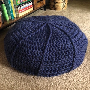 May include: A large, navy blue crocheted floor cushion. The cushion is round and has a textured surface. It is sitting on a beige carpet.