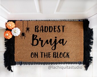May include: A brown coir doormat with black fringe and the text "Baddest Bruja on the Block" in black lettering. Two black spiders are hanging from the top of the mat. The text "@lachiguitasstudio" is at the bottom of the mat.