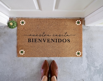 May include: A brown coir doormat with the text "nuestra casita BIENVENIDOS" in black lettering. The doormat has white circles with green centers around the edges.