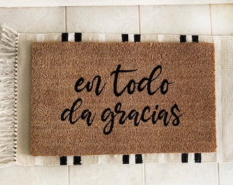 May include: A rectangular brown coir doormat with the phrase "en todo da gracias" in black script. The mat rests on a striped rug with black and white stripes and fringe.