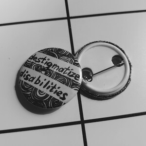 May include: A black and white button with the text "destigmatize disabilities" in a handwritten font. The button is on a white tiled surface.