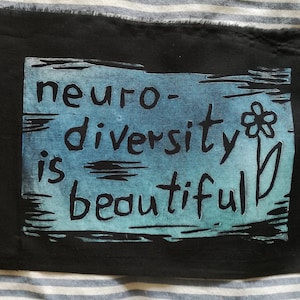 Puede incluir: Parche de tela negro con fondo azul y verde. El texto "neurodiversidad es hermosa" está impreso en negro con una flor y un tallo estilizados.