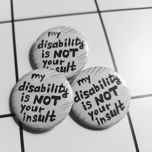 May include: Three white button pins with black text that reads "my disability is NOT your insult".
