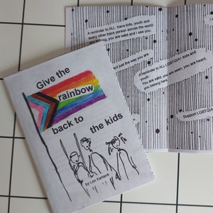 以下が含まれることがあります： 虹の旗と「Give the rainbow back to the kids」というテキストが表紙に書かれた折り畳み式のパンフレット。パンフレットの表紙には、3人の子供たちの白黒のイラストが描かれている。パンフレットの内側のページには、白黒のイラストと「A reminder to ALL LGBTQIA+ kids and youth: You are valid, you are seen, you are heard, you belong here.」と「Support LGBTQIA+ kids and youth.」というテキストが書かれている。