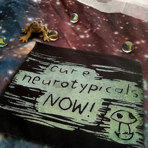 May include: Black fabric patch with white text that reads "cure neurotypicals NOW!" with a green mushroom graphic.