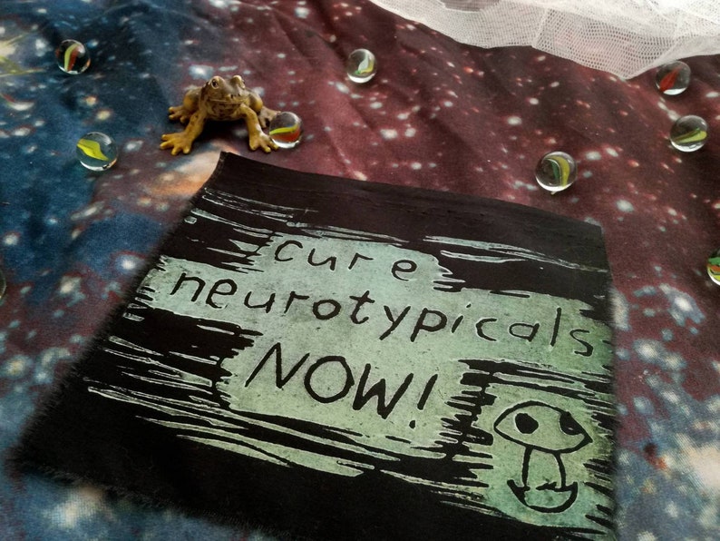 May include: Black fabric patch with white text that reads "cure neurotypicals NOW!" with a green mushroom graphic.