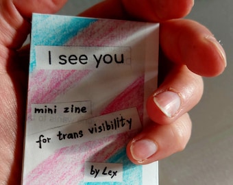 Può includere: Un piccolo zine fatto a mano con una copertina a righe rosa e blu. La copertina ha il testo "Ti vedo" e "mini zine per la visibilità trans di Lex".