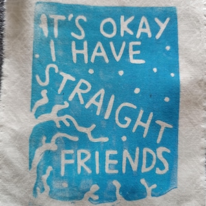 May include: A white fabric square with a blue rectangle in the center. The rectangle has the text "IT'S OKAY I HAVE STRAIGHT FRIENDS" in white. The fabric has a frayed edge.