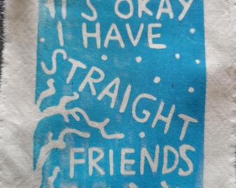 May include: A white fabric square with a blue rectangle in the center. The rectangle has the text "IT'S OKAY I HAVE STRAIGHT FRIENDS" in white. The fabric has a frayed edge.