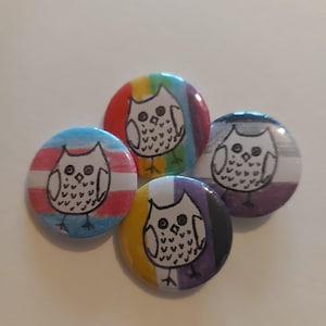 May include: Four round buttons with different pride flag designs. Each button features a black and white drawing of an owl.