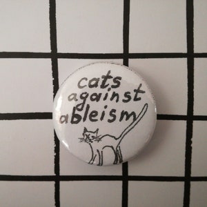 Può includere: Bottone bianco con testo nero e un'illustrazione in bianco e nero di un gatto. Il testo dice "cats against ableism".