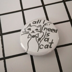 May include: White button with a black and white illustration of a cat wearing a bow tie. The text "all I need is a cat" is written around the cat.