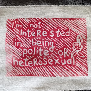 Könnte beinhalten: Ein weißes Stoffquadrat mit einem roten rechteckigen Design. Das Design zeigt den Text "I'm not interested in being polite OR heterosexual" in Weiß, zusammen mit einer floralen Illustration.