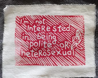 May include: A white fabric square with a red rectangular design. The design features the text "I'm not interested in being polite OR heterosexual" in white, along with a floral illustration.