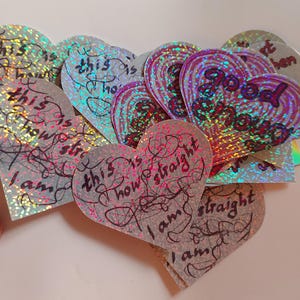 May include: A collection of heart-shaped holographic stickers with the text "This is how straight I am" written in black ink. The stickers are iridescent and have a rainbow effect.