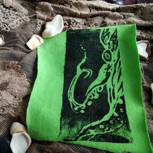 May include: A green fabric patch with a black print of a stylized creature with a long tail and multiple eyes. The creature is surrounded by a black outline.