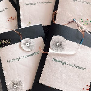 Może przedstawiać: Kilka czarno-białych, ręcznie robionych dzienników z napisem "feelings : activated" wydrukowanym na białej naszywce z tkaniny. Każdy dziennik ma okrągłą, szytą ozdobę i dekoracyjny brokat.