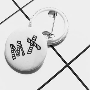 May include: A white button with a black and white striped "MX" design. The button has a metal pin back.