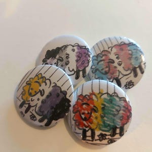 Puede incluir: Cuatro pines redondos con fondo blanco y contornos negros. Cada pin presenta un par de ovejas de dibujos animados con diferentes colores de pelaje, que representan las banderas del orgullo asexual, transgénero, no binario y pansexual.