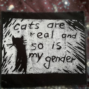 Könnte beinhalten: Schwarz-weißer Grafikaufnäher mit dem Text "cats are real and so is my gender" und einer Katzen-Silhouette. Der Aufnäher hat einen schwarzen Rand und ein strukturiertes, verwittertes Aussehen.