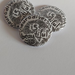 Puede incluir: Tres botones redondos blancos con diseños de patrones en blanco y negro. Los botones tienen el texto "Neurodiversity is beautiful" escrito en ellos.