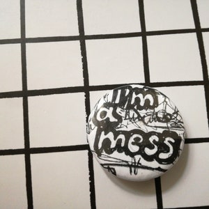 Pode incluir: Um botão branco com texto preto que diz "I'm a mess". O texto está escrito em uma fonte bagunçada e rabiscada.