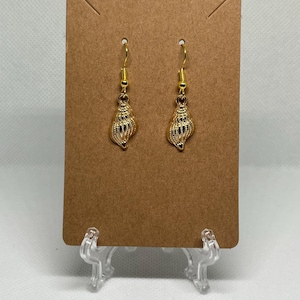 May include: A pair of gold-colored seashell earrings displayed on a brown card. The earrings have a detailed, textured design and are suspended from gold-colored hooks. The background is a plain white surface.