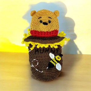 May include: A handmade crochet Winnie the Pooh character emerging from a brown honey pot. The bear is tan with a red shirt, and the pot has a yellow rim and a white and yellow bee applique. The pot has the word "HONEY" on it.