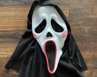 May include: A white plastic Halloween mask with a wide open mouth and red swirl designs on the cheeks. The mask is attached to a black fabric hood.