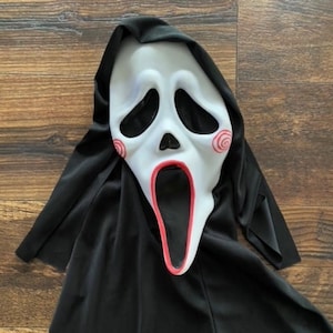 May include: A white plastic Halloween mask with a wide open mouth and red swirl designs on the cheeks. The mask is attached to a black fabric hood.