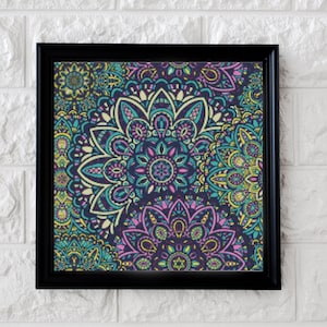 Op de afbeelding: Een ingelijst drukwerk met een kleurrijk mandala-ontwerp met ingewikkelde patronen in tinten blauw, groen, roze en geel. De mandala is geplaatst tegen een donkerblauwe achtergrond.