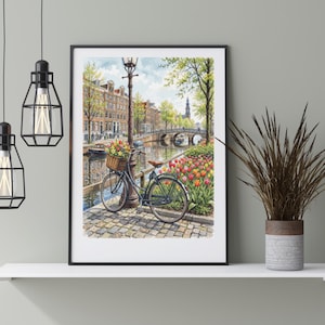 May include: A framed wall art print depicting a black bicycle with a flower-filled basket parked on a cobblestone path beside a canal in Amsterdam. The scene includes a street lamp, colorful tulips, historic brick buildings, and a stone bridge.