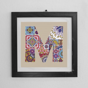 Könnte beinhalten: Ein gerahmter Druck des Buchstabens "M" aus bunten, gemusterten Fliesen. Der Buchstabe befindet sich in einem schwarzen Rahmen vor einer weißen Wand.