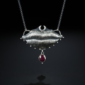 May include: A silver-toned necklace featuring a stylized mouth pendant with textured detailing. A crescent moon shape sits above the mouth, and a teardrop-shaped, deep red gemstone hangs below. The necklace hangs on a delicate chain.