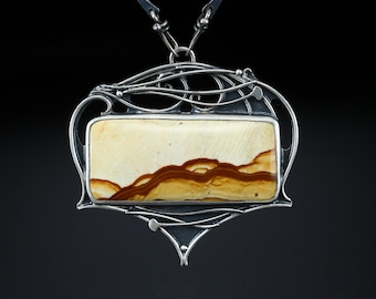 May include: A silver pendant necklace featuring a rectangular stone with a landscape design in shades of cream, brown, and yellow. The stone is set in a decorative silver frame with an Art Nouveau style. The necklace has a dark silver chain.