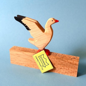 Könnte beinhalten: Ein Holzspielzeug in Form einer Gans mit ausgebreiteten Flügeln, das auf einem Holzblock sitzt. Die Gans hat eine natürliche Holzoberfläche mit schwarzen Flügelspitzen und einem roten Schnabel und Füßen. Ein gelbes Etikett trägt die Aufschrift "Scherbak Wooden Toys".