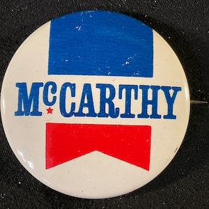 May include: A white button with a blue and red design. The button has the text "McCarthy" in blue letters with a red star.