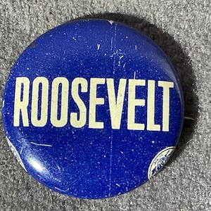 May include: A round, blue button with the word "ROOSEVELT" in large, white capital letters. The button has a slightly worn appearance, suggesting it is vintage. A small, circular design is visible near the edge.