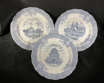 May include: Three white ceramic plates with scalloped edges and blue transferware patterns. Each plate features a central architectural illustration and text, surrounded by a detailed geometric and floral border in blue and white.