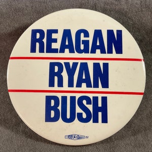May include: A white campaign button with the names "REAGAN", "RYAN", and "BUSH" in blue block letters. The names are separated by red horizontal lines. The button has a vintage look.