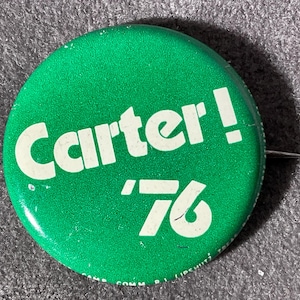 May include: A green campaign button with the word "Carter!" in white, along with the number "'76". The button has a pin on the back and is likely a political collectible.