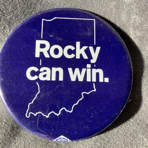 May include: A round, dark blue button with the text "Rocky can win." in white. The button features a white outline of the state of Indiana. A small white design is at the bottom.
